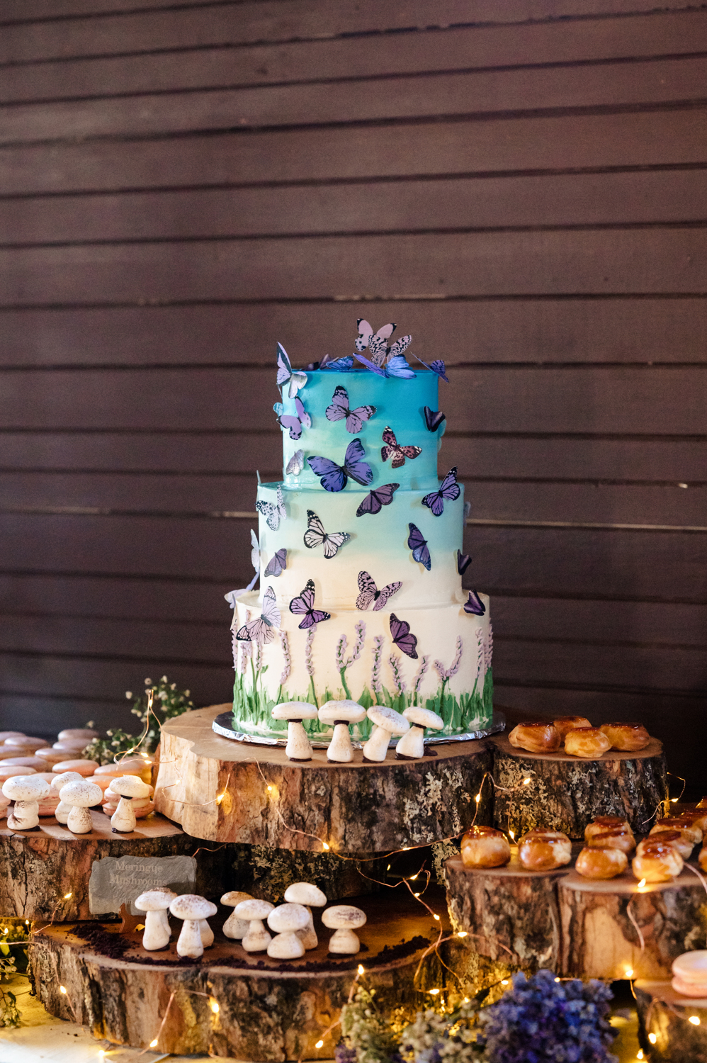Fanciful wedding dessert spread by The Goose Chase Cake Design, with a 3-tiered ombre butterfly cake, mushroom-shaped meringues, macarons, and cream puffs on rustic wood cookies with fairy lights and florals.